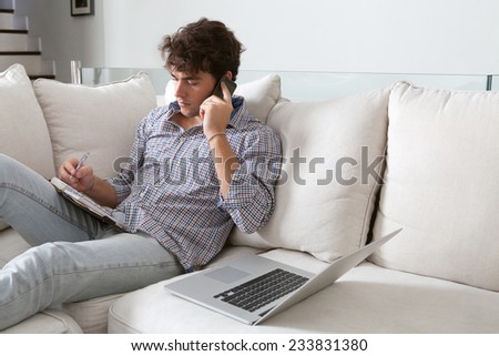 Young attractive business man sitting on a white sofa in his home living room, making a call on his smartphone and using a laptop computer. Professional businessman working from home, sitting on sofa.