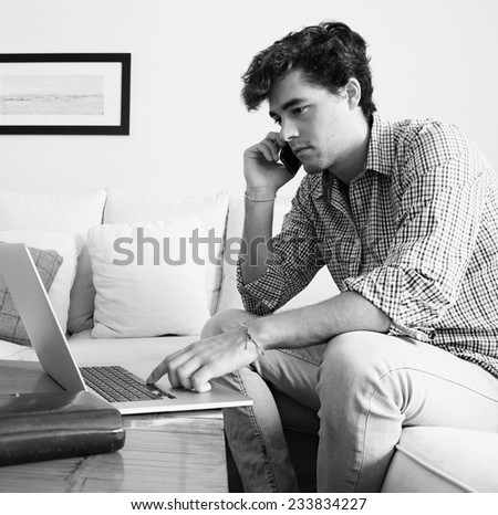 Black and white portrait of professional businessman sitting on sofa in home living room, using a laptop computer and smartphone working from home. Business man on phone conversation in home setting.