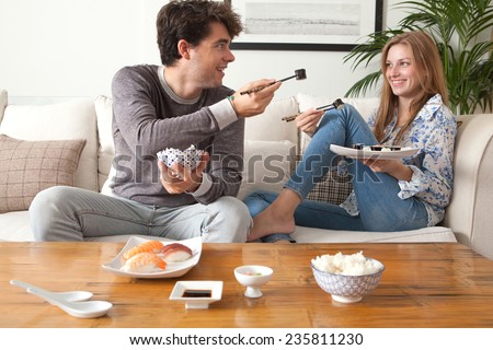 Attractive young couple having a night in at home and enjoying eating japanese take away food. Couple relaxing on home sofa eating exotic food with man offering food in a home interior.