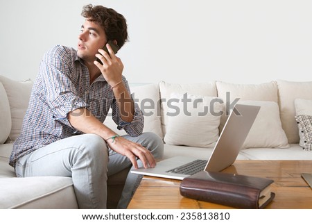 Working business man having a phone conversation on a smart phone and using a laptop working from home office. Young professional man using technology in a home living room, interior.