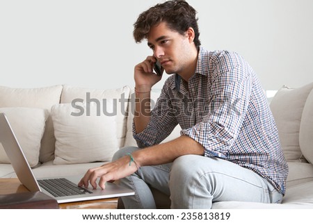 Attractive young professional businessman sitting on white sofa in home living room, using a laptop computer and smartphone working from home. Business man on phone conversation in home setting.