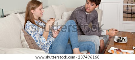 Panoramic view of couple enjoying eating Japanese sushi and maki food at home, sitting on a white couch in a home living room, sharing food and having a good time, smiling together. Eating fresh food.