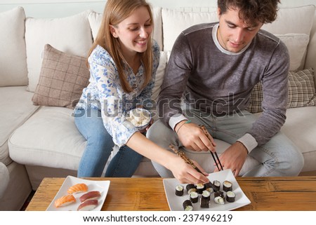 Portrait of attractive couple enjoying eating Japanese sushi and maki food at home, sitting on a white couch in a home living room, sharing food and smiling together. Eating fresh food.