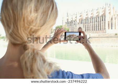 Rear portrait view of a young tourist woman on vacation, taking pictures with her smart phone touch screen technology of a cathedral monument while sightseeing in a destination city, outdoors.