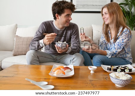 Portrait of attractive young couple having a night in at home and enjoying eating japanese take away food together. Couple relaxing on home sofa eating exotic food in a stylish home interior.