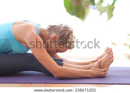 Side view of a mature professional woman exercising and stretching her body doing flexibility exercises and bending her back, sitting on a yoga mat indoors. Senior woman fit and healthy lifestyle.