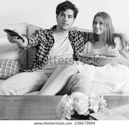 Black and white view of young couple sitting together on white sofa at home using a control remote to watch television, smiling eating pop corn together. Home lifestyle and entertainment technology.