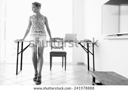 Black and white view of a mature professional business woman working from home and sitting on the edge of her work desk with a plasma tv blank screen. Home and office technology.