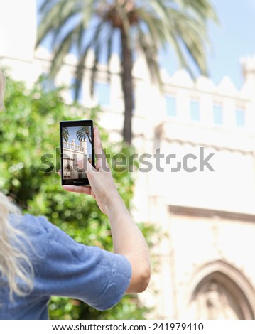 Rear portrait view of a young tourist girl holding up a smartphone device to take pictures of a monument while visiting a destination city on holiday. Vacation travel and technology networking.
