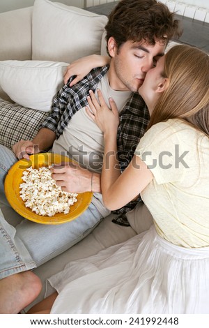 Close up portrait of young couple sitting together on a sofa at home watching television, eating popcorn and kissing on the lips, being romantic. Home and relationships lifestyle and entertainment.