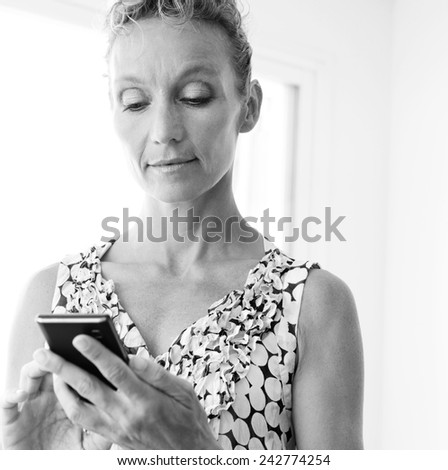 Black and white portrait of an attractive mature professional business woman in an office space using a smartphone device to connect on line, workplace interior. Senior businesswoman using technology.