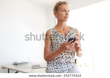 Thoughtful powerful mature professional business woman in an office space, sitting on her desk and using a smartphone, workplace interior. Senior businesswoman using technology.