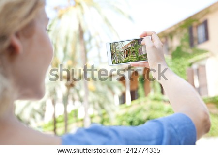 Rear view of a young tourist girl holding up a smartphone device to point and take pictures of a monument while visiting a destination city on holiday. Vacation travel and technology networking.