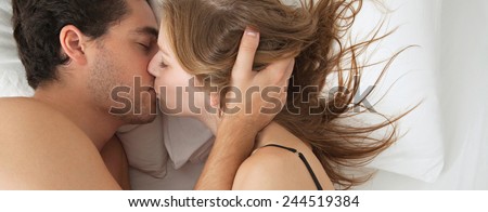 Overhead panoramic portrait of a young couple caressing laying in bed together being romantic hugging and kissing. Couple in a relationship caressing in a white bed, home sex interior lifestyle.