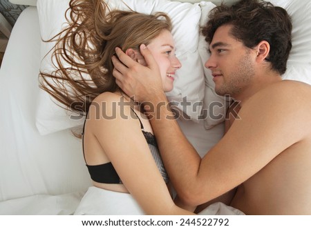 Overhead side portrait of an attractive young couple caressing laying in bed together in lingerie being romantic. Couple in a relationship caressing after sex in a white bed, home interior lifestyle.