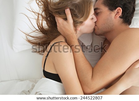 Overhead close up portrait of a young couple caressing laying in bed together in lingerie being romantic hugging and kissing. Couple in a relationship having sex in white bed, home interior lifestyle.