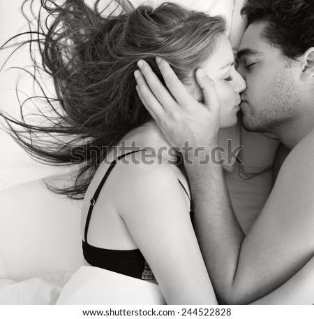 Overhead black and white portrait of a young couple caressing laying in bed together being romantic hugging and kissing. Couple in a relationship having sex in a white bed, home interior lifestyle.
