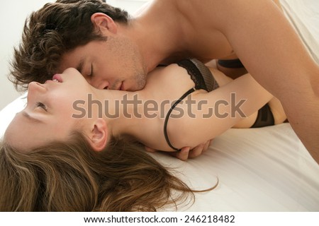 Close up portrait of young attractive romantic couple hugging and kissing, laying down on a white bed, having sex and being loving with each other. Love and relationships lifestyle, interior bedroom.