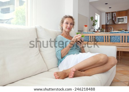Mature woman sitting and relaxing on a white couch at home while eating a small green salad, home interior. Senior woman eating healthy food. Wellness and well being indoors. Lifestyle, smiling.