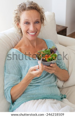 Mature woman sitting and relaxing on a white couch at home while eating a small green salad, home interior. Senior woman eating healthy food. Wellness and well being indoors. Lifestyle, smiling.