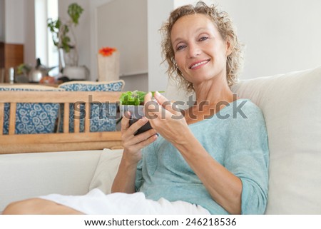 Close up portrait of mature woman sitting on a couch at home while eating a small green salad, home interior. Senior woman eating healthy food.Wellness and well being indoors. Lifestyle, smiling.
