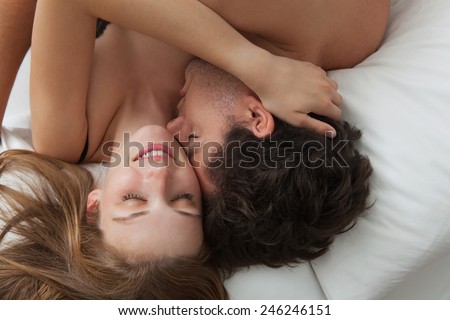 Overhead close up portrait of a young romantic couple hugging and kissing, laying down on a white bed, having sex and loving each other. Love and relationships lifestyle, interior bedroom.