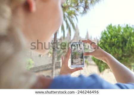 Rear portrait view of a young tourist girl holding up a smartphone device and take pictures of a monument while visiting a destination city on holiday. Vacation travel and technology networking.