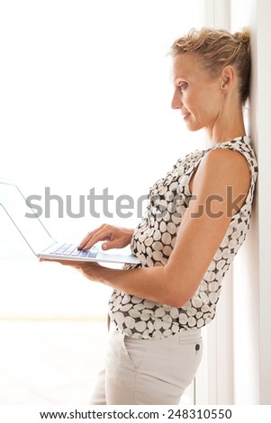 Side view of a mature professional woman using a laptop computer while leaning on an office wall next to a bright window with white space. Business and lifestyle technology, interior.