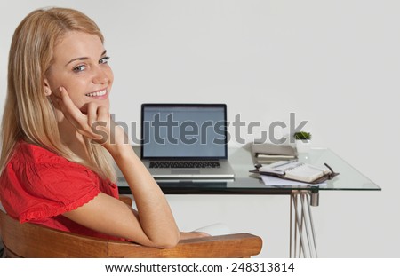 Portrait of an attractive young student using laptop technology at home to study and do her homework, sitting at her home desk with a computer and paperwork. Young professional smiling indoors.