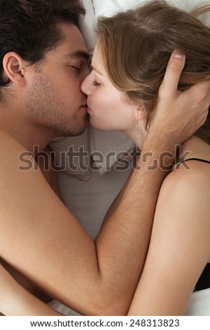 Overhead close up portrait of a young couple caressing laying in bed in lingerie being romantic hugging and kissing. Couple caressing while having sex in a white bed, home interior lifestyle.