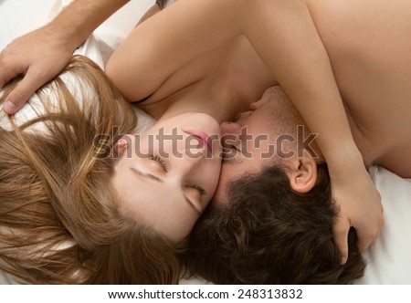 Overhead close up portrait of a young couple caressing laying in bed together, hugging with closed eyes. Couple in a relationship caressing while having sex in a white bed, home interior lifestyle.