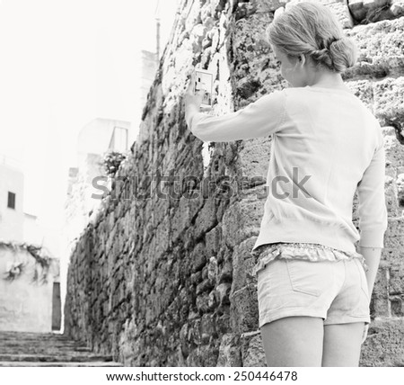 Black and white rear view of a young tourist woman in a picturesque stone street, holding up and using a smartphone device to take pictures and network while on a sunny holiday. Travel and technology.