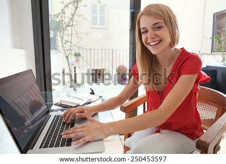 Portrait of a young professional woman smiling in her home office space, typing and joyfully looking at the camera. Student girl using a laptop computer at home. Lifestyle and technology, interior.