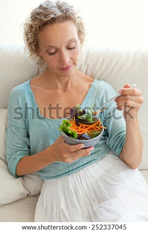 Beautiful mature woman sitting and relaxing on a white sofa at home while eating a small green salad, home interior. Senior woman eating healthy food, wellness and well being indoors. Lifestyle.