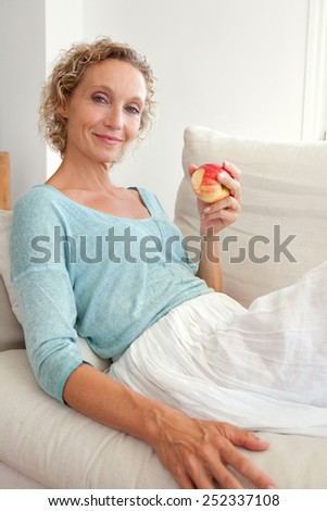 Portrait of a mature healthy woman eating a red apple at home, relaxing on a white sofa in a home living room against a white wall, smiling indoors. Healthy eating and well being lifestyle, interior.