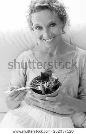 Black and white portrait of a smiling mature woman on a white sofa at home, eating a small green salad, home interior. Senior woman eating healthy food, wellness and well being indoors. Lifestyle.