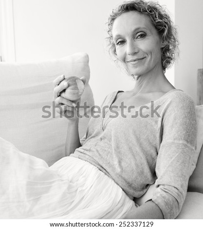 Black and white portrait of a mature healthy woman eating an apple at home while relaxing on a white sofa against a white wall, smiling indoors. Healthy eating and well being lifestyle, interior.