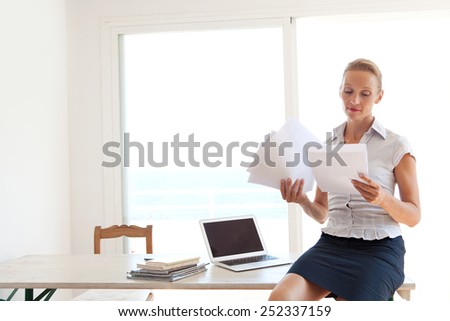 Confident senior business woman sitting on her home office desk handling and reading paperwork, working with a laptop computer, interior. Professional businesswoman using technology.