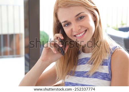Portrait of a young teenager girl joyfully smiling at home using a smartphone mobile phone to have a phone conversation next to a large window. Young student lifestyle and technology, interior.