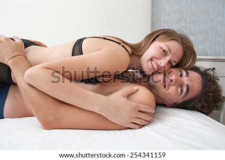 Side profile view of young lovers couple laying down together on a bed, with the girl laying on top of the man being playful and joyful indoors. Relationships lifestyle and sex life. Romance living.