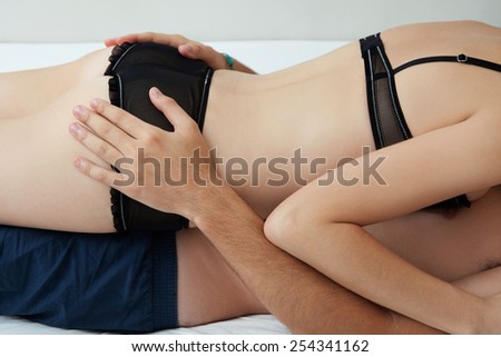 Side profile of a young lovers couple body section laying down together on a white bed, with the girl laying on top of the man being playful indoors. Relationships lifestyle and sex life.