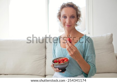 Close up portrait of a mature woman smiling and eating red strawberries at home, relaxing on a white sofa in a home living room, indoors. Healthy eating and well being lifestyle, interior.
