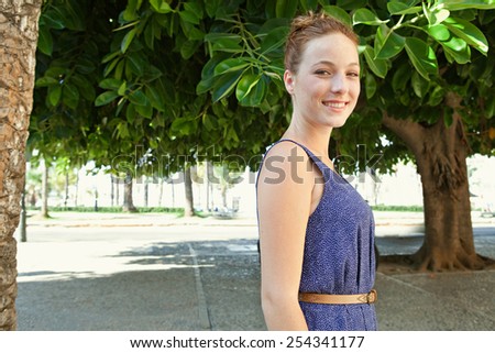 Side portrait of a beautiful young tourist woman visiting a green tree garden park while on holiday, outdoors. Travel and lifestyle. Attractive woman walking and looking at camera, smiling.