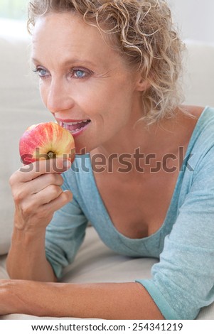 Close up portrait of a mature healthy woman biting and eating a red apple at home while relaxing on a white sofa in a home living room, indoors. Healthy eating and well being lifestyle, interior.