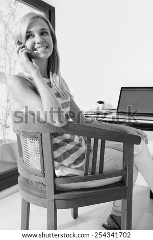 Black and white portrait of teenager girl student at her work desk at home, using smartphone technology, and a laptop computer to work and study. Professional woman with mobile phone indoors, smiling.