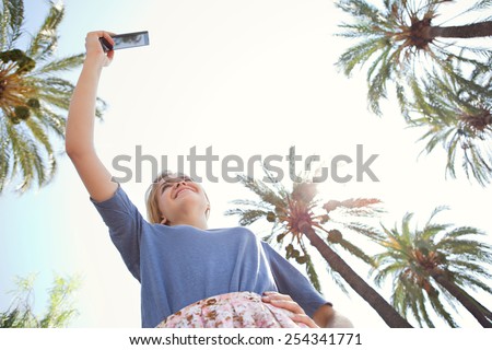 Under view of young tourist woman raising her arm holding and pointing a smartphone device to take pictures of palm trees on a sunny blue sky on summer holiday, outdoors. Travel technology lifestyle.