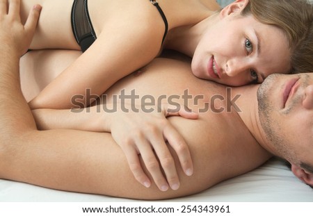 Portrait of attractive young couple hugging and caressing in a hotel bed being in love, wearing a black bra, relaxing in a bedroom with white linen. Love, passion and romance lifestyle, home interior.