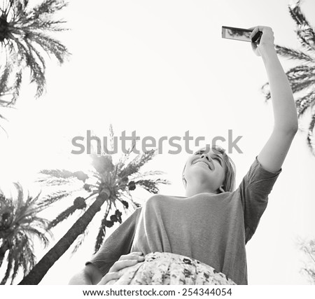 Black and white low view of a young tourist woman raising her arm pointing a smartphone device to take pictures of palm trees on a sunny sky on holiday, outdoors. Travel and technology lifestyle.