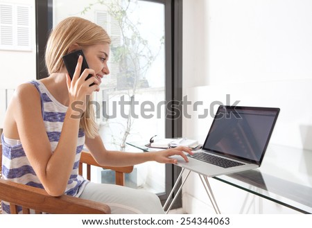 Young beautiful woman working at home office space using a smartphone mobile phone to have a call conversation. Student using a laptop computer at home work desk. Lifestyle and technology, interior.