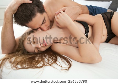 Close up portrait of attractive young lovers couple together in bed with man kissing woman on neck, wearing sexy lingerie in a hotel room, interior. Romance and sex lifestyle indoors. Relationships.